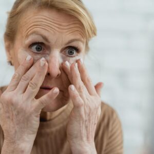 Crop focused mature female applying moisturizing nourishing eye patches and looking away in light bathroom